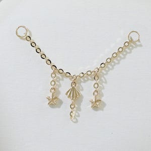 Könnte beinhalten: Eine zarte goldfarbene Kette mit drei Anhängern: einer Muschel und zwei Sternen. Die Kette ist so konzipiert, dass sie mit zwei kleinen Schlaufen an jedem Ende an einer Oberfläche befestigt werden kann. Der Schmuck wird vor weißem Hintergrund präsentiert.