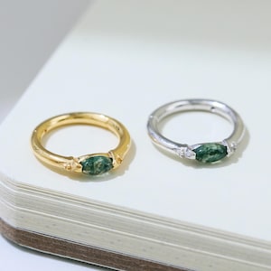 Puede incluir: Dos anillos, uno dorado y otro plateado, cada uno con una piedra preciosa verde ovalada. El anillo dorado tiene una banda delicada y pequeñas piedras de acento. El anillo plateado tiene un diseño similar. Ambos anillos están sobre una superficie blanca.