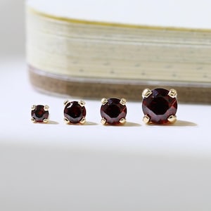 May include: Four gold stud earrings with round, deep red gemstones of varying sizes are arranged in a row. The earrings are set against a white surface with a blurred notebook in the background. The gemstones have a rich, dark colour.