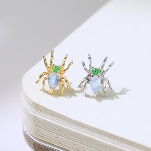 May include: Gold and silver spider-shaped stud earrings with green and blue accents. The earrings are displayed on a white surface. A small gold dinosaur-shaped earring is in the upper left corner.