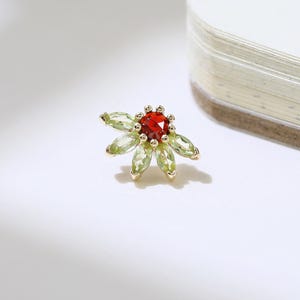 Op de afbeelding: Een goudkleurige bloemenstud-oorbel met een rode middensteen en groene, bloemblad-vormige stenen. De oorbel is geplaatst tegen een witte achtergrond met een notitieboek. Sieraden.