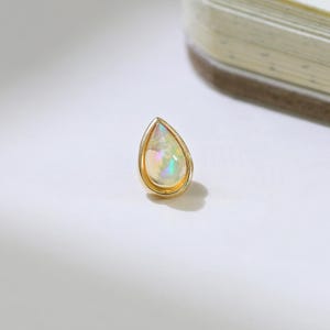 Pode incluir: Um brinco de pressão em forma de gota, com uma moldura dourada e uma pedra de opala brilhante. A opala exibe uma gama de cores, incluindo branco, amarelo e verde. O brinco está sobre um fundo branco.