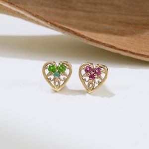 May include: A pair of gold heart-shaped stud earrings. One earring features green gemstones, the other features red gemstones. The earrings are on a white surface with a wooden element in the background.