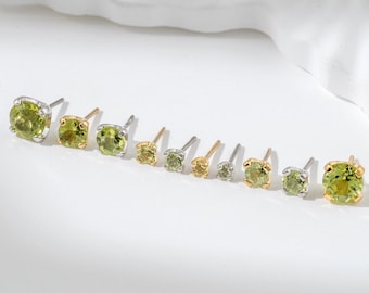 Könnte beinhalten: Eine Auswahl an Peridot-Ohrsteckern in verschiedenen Größen, präsentiert auf einer weißen Oberfläche. Die Ohrringe haben runde, hellgrüne Edelsteine, die in Gold- und Silberfassungen gefasst sind. Die Ohrringe sind in einer Reihe angeordnet, um ihre unterschiedlichen Größen hervorzuheben.