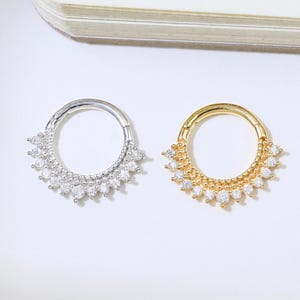 May include: Two septum rings, one silver and one gold, each featuring a row of clear gemstones. The rings have a sunburst design, adding a touch of elegance. The jewellery is displayed on a white surface. The gemstones sparkle.