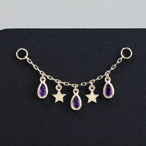May include: A gold-coloured chain featuring purple teardrop and star charms. The chain is attached to a black surface. The charms are evenly spaced, creating a delicate and elegant design.