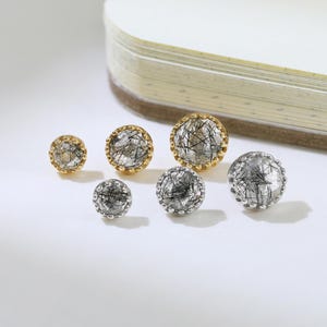 May include: A collection of gemstone stud earrings in various sizes. The earrings feature a gold or silver setting with a clear, round stone containing black inclusions. The earrings are displayed on a white surface next to a notebook.