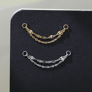 May include: Two delicate chain earrings, one gold and one silver, are displayed against a black background. Each earring features two chains connected by small rings, creating a graceful, curved shape. The earrings are simple and elegant.