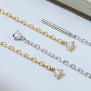 以下が含まれることがあります： 繊細なイヤリングのペアのクローズアップ。イヤリングは、端に小さな透明な宝石が付いたチェーンのデザインが特徴です。一部は金色のトーンで、その他は銀色のトーンです。イヤリングは白い表面に表示されます。