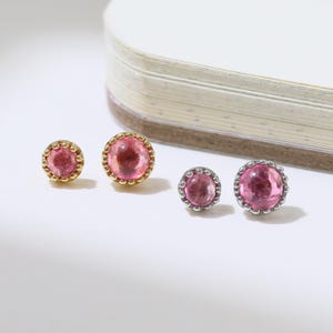 Può includere: Quattro paia di orecchini a lobo con pietre preziose rosa. Due paia hanno montature color oro e gli altri due color argento. Le pietre preziose sono di un rosa vibrante e gli orecchini sono esposti su una superficie bianca.