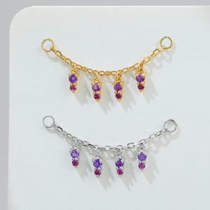 May include: Two necklaces, one gold-coloured and one silver-coloured, each with a series of dangling charms. The charms alternate between purple and red gemstones. The necklaces are displayed on a white surface, creating a striking contrast.