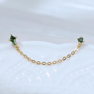 May include: A delicate gold chain earring with two green gemstone accents. One gemstone is a diamond shape, and the other is a small square. The earring is on a white, textured surface.