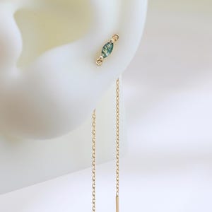 May include: A close-up of a gold earring set. The earring features a small, green gemstone stud and a delicate chain that hangs down with a gold bar at the end. The earring is displayed on a white ear model.