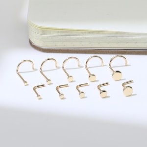May include: A selection of gold-coloured nose rings in different styles, including L-shaped and screw designs, with small, round, flat studs. The jewellery is displayed on a white surface next to a notebook.