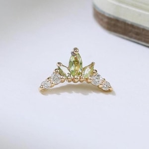 May include: A gold-colored, V-shaped earring with a central light green gemstone and surrounding clear gemstones. The earring has a delicate, crown-like design, perfect for ear piercings.
