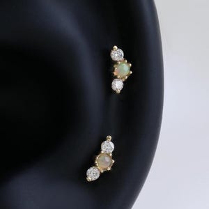 May include: Two gold-toned earrings, each featuring a cluster of three gemstones. The earrings are set with a central opal-like stone, flanked by smaller, clear crystals. The earrings are displayed on a black ear model.