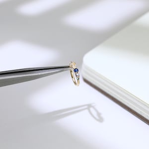 May include: A small, gold ring featuring a row of colourful gemstones. The ring is held by tweezers, highlighting its intricate design. The gemstones include blue, white, and other hues. The ring is likely a piece of jewellery.