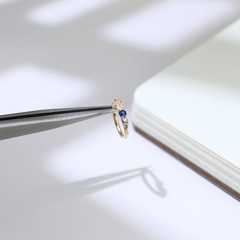 May include: A small, gold ring featuring a row of colourful gemstones. The ring is held by tweezers, highlighting its intricate design. The gemstones include blue, white, and other hues. The ring is likely a piece of jewellery.