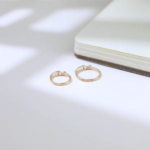 May include: Two gold rings with small, dark gemstones are shown on a white surface. The rings have a delicate design, with the gemstones set in a row on the top. A notebook is partially visible in the background.