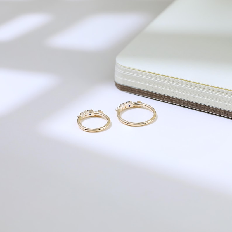 May include: Two gold rings with small, dark gemstones are shown on a white surface. The rings have a delicate design, with the gemstones set in a row on the top. A notebook is partially visible in the background.