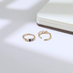 May include: Two gold rings with gemstone detailing. One ring is closed, adorned with a dark blue gemstone and smaller purple and green stones. The other ring is open, displaying similar gemstone features. The rings are on a white surface.