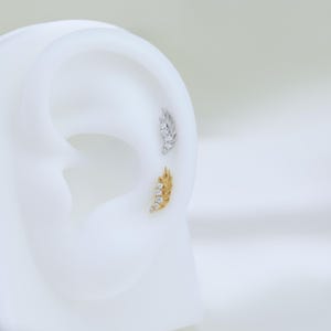 May include: Two ear piercings, one gold and one silver, each shaped like a feather. The gold feather has four clear stones, and the silver feather has three. The earrings are displayed on a white ear model.