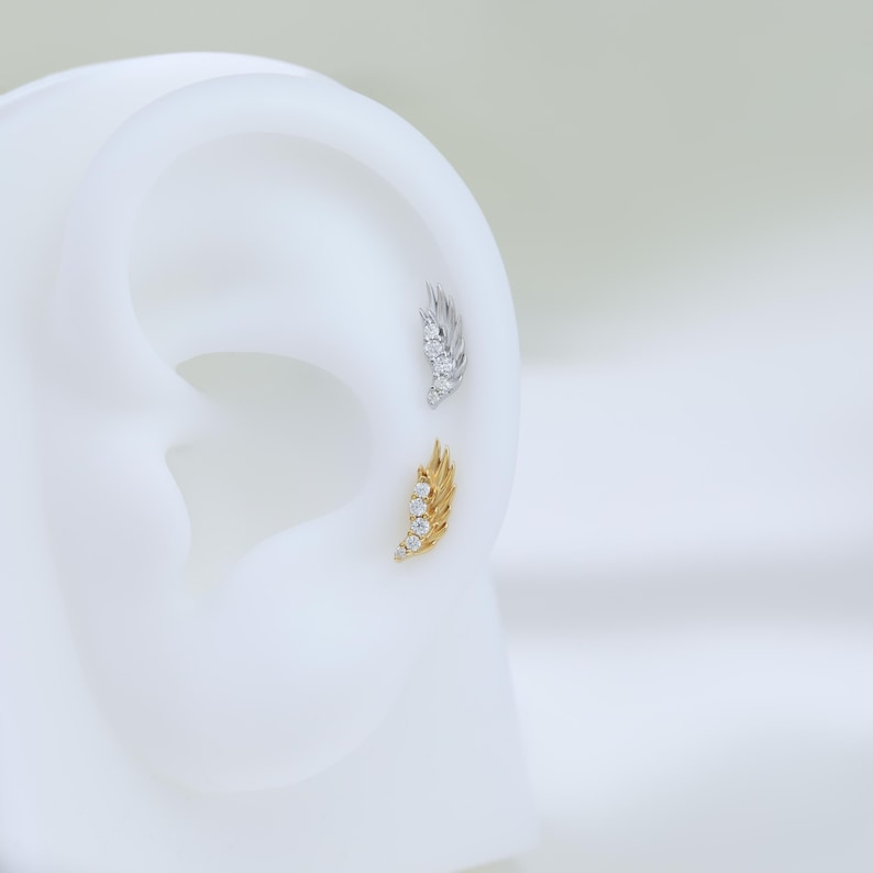 May include: Two ear piercings, one gold and one silver, each shaped like a feather. The gold feather has four clear stones, and the silver feather has three. The earrings are displayed on a white ear model.