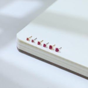 May include: A collection of stud earrings with vibrant red gemstones. The earrings are displayed in a row, showcasing variations in metal colors, including gold and silver. The gemstones are round and set in a classic prong setting, offering a timeless jewelry design.