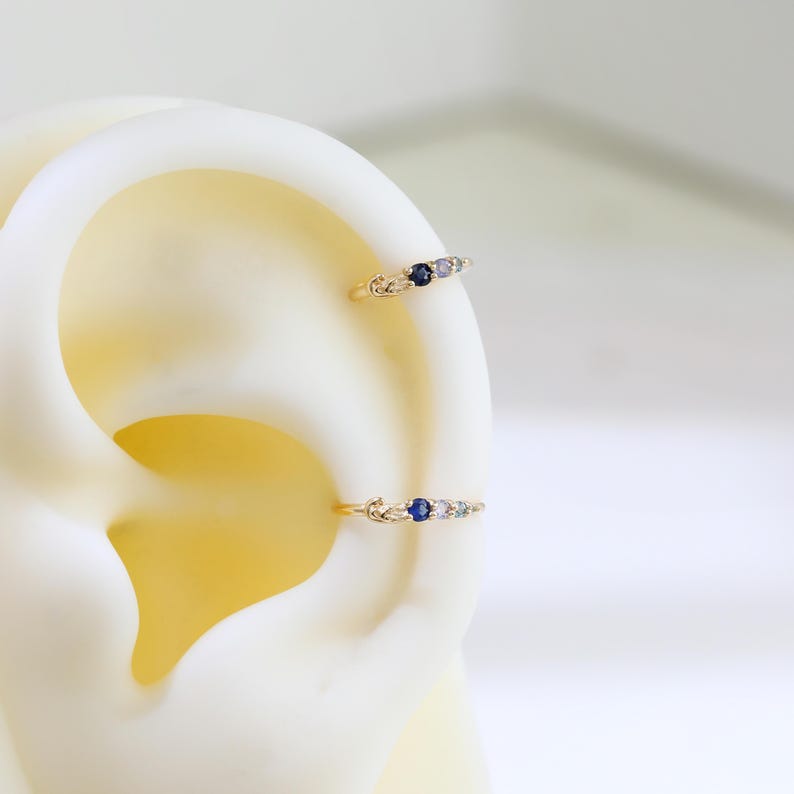 May include: Two gold-coloured hoop earrings, each featuring three small gemstones. The gemstones are a mix of blue, purple, and clear, set in a delicate design. The earrings are displayed on a white ear model.