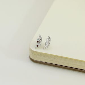 May include: A pair of silver-toned wing-shaped earrings. One earring features small red and blue gemstones, while the other has clear gemstones. The earrings are displayed on a cream-colored surface, suggesting a jewelry or accessory theme.