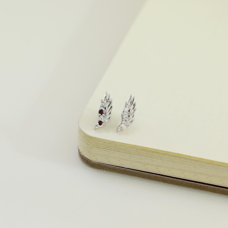 May include: A pair of silver-toned wing-shaped earrings. One earring features small red and blue gemstones, while the other has clear gemstones. The earrings are displayed on a cream-colored surface, suggesting a jewelry or accessory theme.