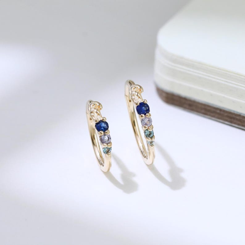 May include: A pair of gold hoop earrings, each embellished with a sequence of colourful gemstones. The earrings showcase a mix of blue, purple, and green stones, creating a vibrant design. The earrings are set against a white background.