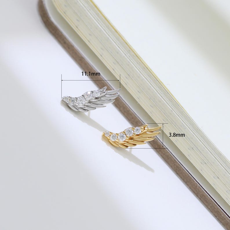 May include: Two wing-shaped earrings, one silver-toned and one gold-toned, each adorned with clear gemstones. The silver earring measures 0.44 inches in length, and the gold earring is 0.15 inches long. The earrings are displayed on a light-colored surface.