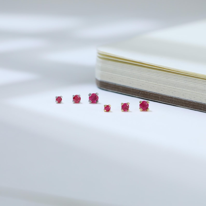 May include: A collection of small, round ruby earrings in varying sizes. The earrings are set in silver and gold settings, displayed on a white surface next to a partially visible notebook. The rubies are a vibrant red color.