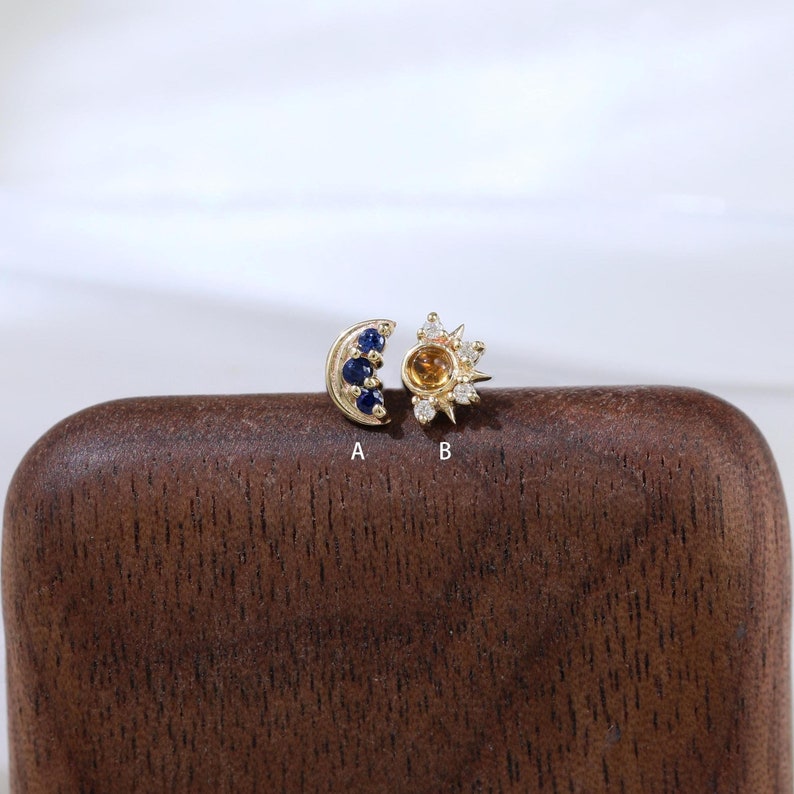 May include: Two gold earrings with gemstones. The earring on the left is a crescent moon shape with blue gemstones. The earring on the right is a sun shape with a yellow gemstone in the center and white gemstones around it. The earrings are on a brown wooden surface. A and B are labeled below the earrings.