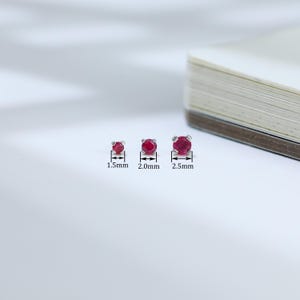 May include: Three ruby stud earrings of varying sizes are displayed. The smallest is 1.5mm, the middle is 2.0mm, and the largest is 2.5mm. Each earring features a round, vibrant red gemstone set in a silver or white metal setting.