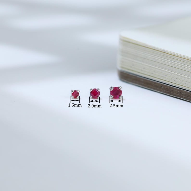 May include: Three ruby stud earrings of varying sizes are displayed. The smallest is 1.5mm, the middle is 2.0mm, and the largest is 2.5mm. Each earring features a round, vibrant red gemstone set in a silver or white metal setting.