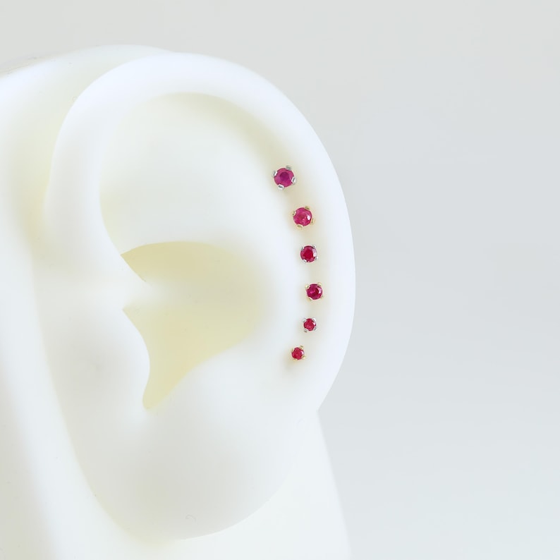 May include: A series of seven small, round, ruby-colored earrings are displayed in a descending size order along the curve of a white ear model. The earrings are set in a gold-colored metal.