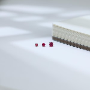 May include: Three silver stud earrings with round, vibrant red gemstones of varying sizes are arranged on a white surface. The earrings are set against a soft, blurred background, suggesting a focus on the jewelry.