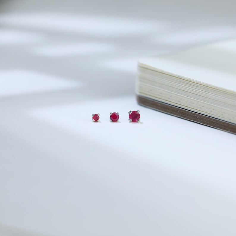 May include: Three silver stud earrings with round, vibrant red gemstones of varying sizes are arranged on a white surface. The earrings are set against a soft, blurred background, suggesting a focus on the jewelry.