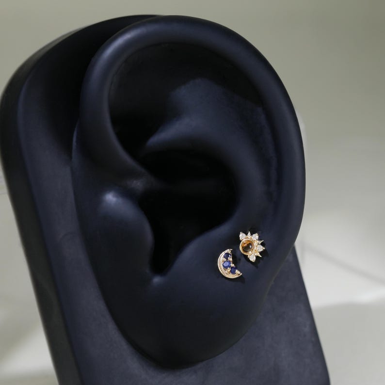 May include: A close-up of a person's ear with two gold earrings. The earring on the top is a sun with a yellow center and white diamonds. The earring on the bottom is a crescent moon with blue sapphires.