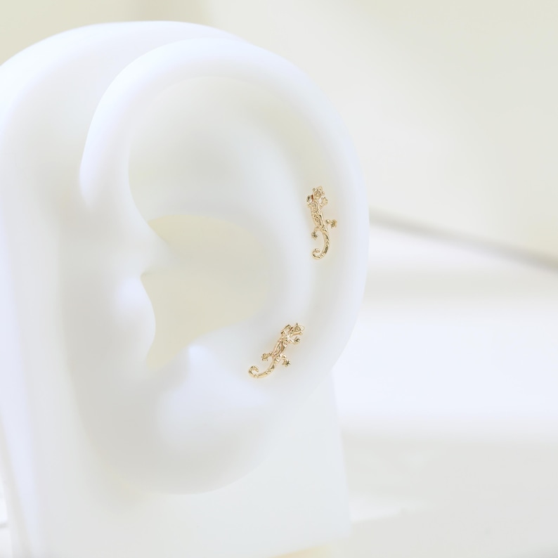 May include: A pair of gold-colored lizard-shaped stud earrings. The earrings are small and detailed, with a textured surface. The lizards are positioned on a white surface, suggesting they are being displayed for sale. The earrings are likely made of metal.