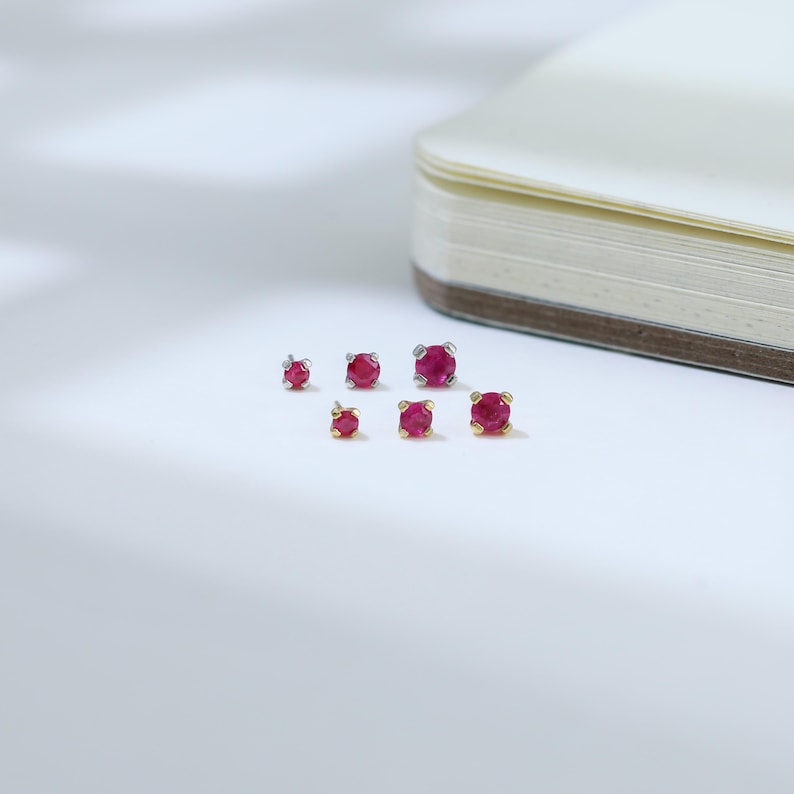 May include: A collection of ruby stud earrings in various sizes. The earrings feature vibrant red gemstones set in gold and silver settings. The studs are displayed on a white surface, with a notebook in the background.