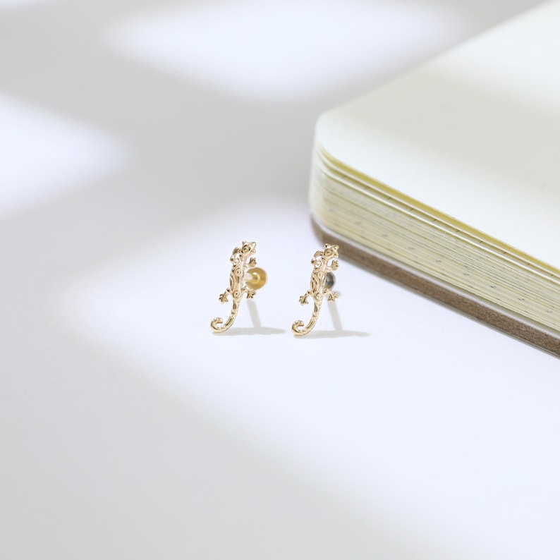 May include: A pair of gold-colored lizard-shaped stud earrings. The earrings are detailed with textured scales and small, round accents. The earrings are displayed on a white surface.