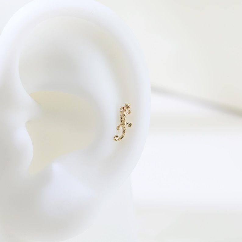 May include: A gold-colored lizard-shaped earring. The earring is a small, detailed representation of a lizard, with its legs and tail curved. The earring is set against a white background, highlighting its intricate design and color.