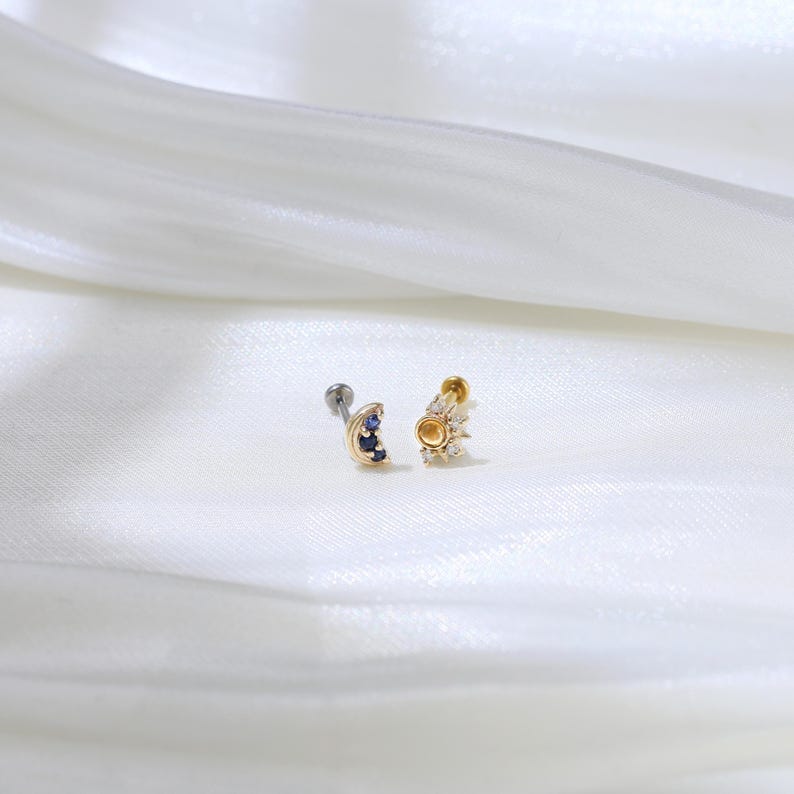 May include: Two gold body jewelry pieces, one with a crescent moon design with blue gemstones and the other with a sun design with clear gemstones.