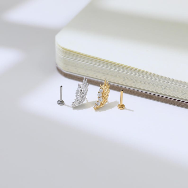 May include: Two wing-shaped earrings, one silver and one gold, each adorned with three clear gemstones. Two additional earring posts are also visible. The earrings are displayed on a white surface next to a partially visible open book.