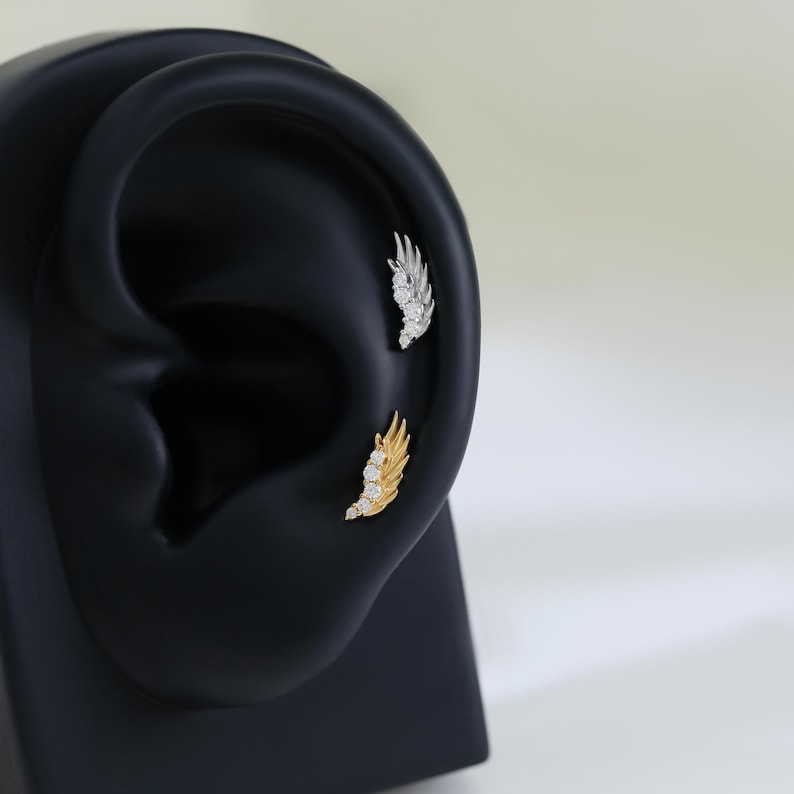 May include: A pair of ear piercings, one gold and one silver, each shaped like a wing. The gold wing has three clear stones, and the silver wing has three clear stones. The earrings are displayed on a black ear model.