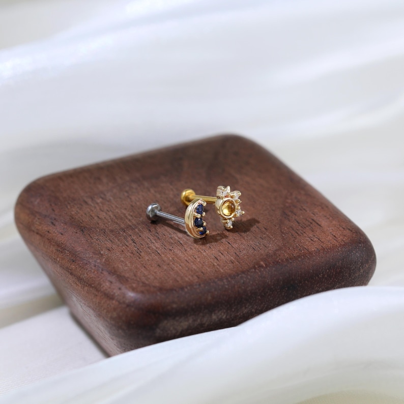 May include: Two gold nose rings with clear and blue gemstones. The ring on the left has a crescent moon shape with blue gemstones. The ring on the right has a flower shape with clear gemstones.