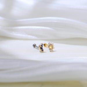 May include: Two gold body jewelry pieces, one with a crescent moon shape with blue gemstones and the other with a sun shape with clear gemstones.